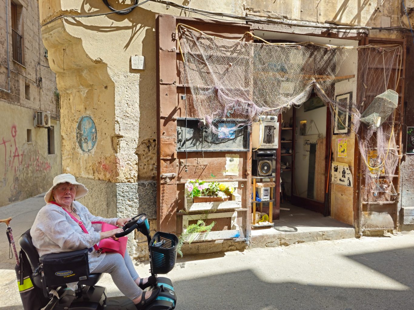 Italy 2024 80 year old lady wearing a hat, sitting on a mobility scooter with an old building in the background where there is a shop visible.